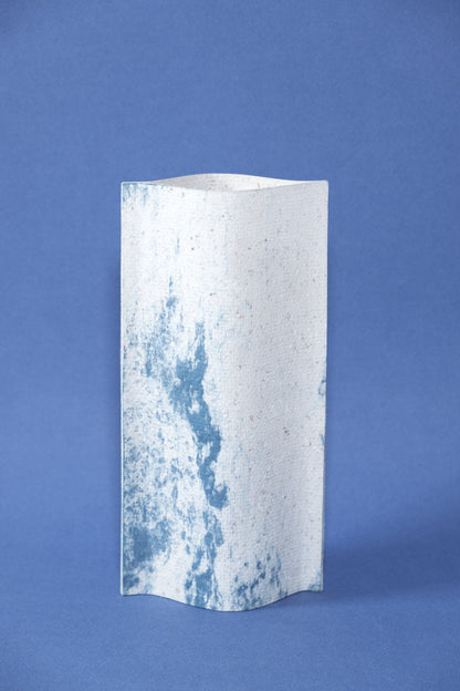 Un vase de profil rectangulaire sur fond bleu foncé. Deux feuilles de carton-pierre sont collées à leurs extrémités et ouvertes en leur centre pour y laisser passer un contenant étanche. Le vase arbore un effet marbré avec des veines couleur bleu marine sur fond blanc. Chaque vase a un marbré unique.