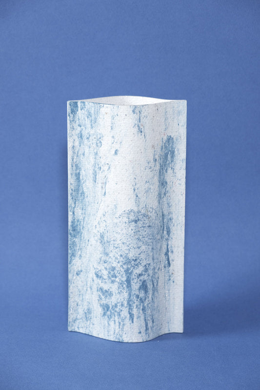 Un vase de profil rectangulaire sur fond bleu foncé. Deux feuilles de carton-pierre sont collées à leurs extrémités et ouvertes en leur centre pour y laisser passer un contenant étanche. Le vase arbore un effet marbré avec des veines couleur bleu marine sur fond blanc. Chaque vase a un marbré unique.