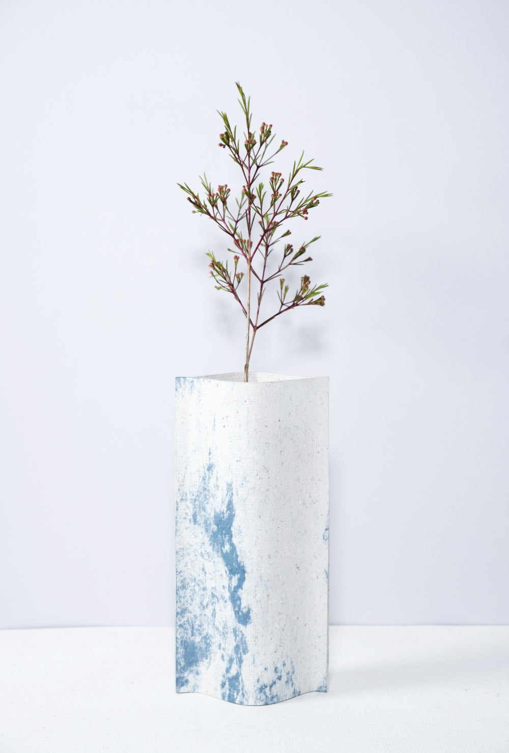 Un vase de profil rectangulaire sur fond blanc. Une branche y est soigneusement disposée. Deux feuilles de carton-pierre sont collées à leurs extrémités et ouvertes en leur centre pour y laisser passer un contenant étanche. Le vase arbore un effet marbré avec des veines couleur bleu sur fond blanc. Chaque vase a un marbré unique.