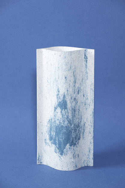 Un vase de profil rectangulaire sur fond bleu foncé. Deux feuilles de carton-pierre sont collées à leurs extrémités et ouvertes en leur centre pour y laisser passer un contenant étanche. Le vase arbore un effet marbré avec des veines couleur bleu marine sur fond blanc. Chaque vase a un marbré unique.