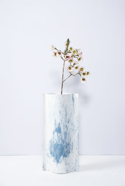 Un vase de profil rectangulaire sur fond blanc. Une branche y est soigneusement disposée. Deux feuilles de carton-pierre sont collées à leurs extrémités et ouvertes en leur centre pour y laisser passer un contenant étanche. Le vase arbore un effet marbré avec des veines couleur bleu sur fond blanc. Chaque vase a un marbré unique.