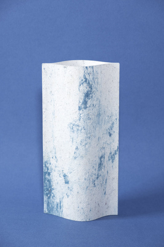 Un vase de profil rectangulaire sur fond bleu foncé. Deux feuilles de carton-pierre sont collées à leurs extrémités et ouvertes en leur centre pour y laisser passer un contenant étanche. Le vase arbore un effet marbré avec des veines couleur bleu marine sur fond blanc. Chaque vase a un marbré unique.