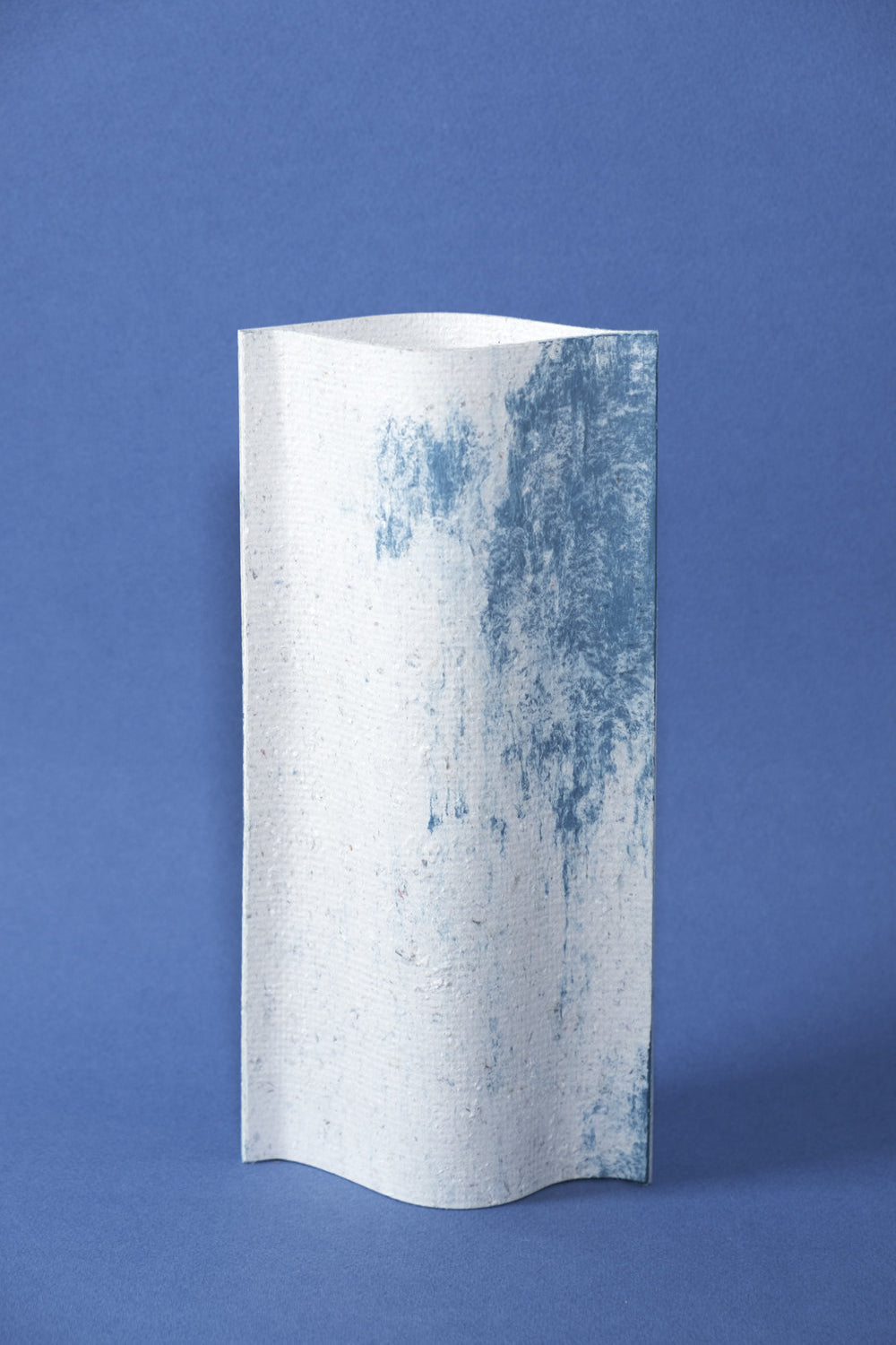 Un vase de profil rectangulaire sur fond bleu foncé. Deux feuilles de carton-pierre sont collées à leurs extrémités et ouvertes en leur centre pour y laisser passer un contenant étanche. Le vase arbore un effet marbré avec des veines couleur bleu marine sur fond blanc. Chaque vase a un marbré unique.