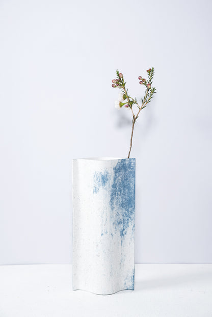 Un vase de profil rectangulaire sur fond blanc. Une branche y est soigneusement disposée. Deux feuilles de carton-pierre sont collées à leurs extrémités et ouvertes en leur centre pour y laisser passer un contenant étanche. Le vase arbore un effet marbré avec des veines couleur bleu sur fond blanc. Chaque vase a un marbré unique.