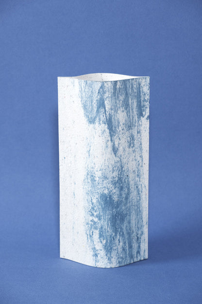 Un vase de profil rectangulaire sur fond bleu foncé. Deux feuilles de carton-pierre sont collées à leurs extrémités et ouvertes en leur centre pour y laisser passer un contenant étanche. Le vase arbore un effet marbré avec des veines couleur bleu marine sur fond blanc. Chaque vase a un marbré unique.