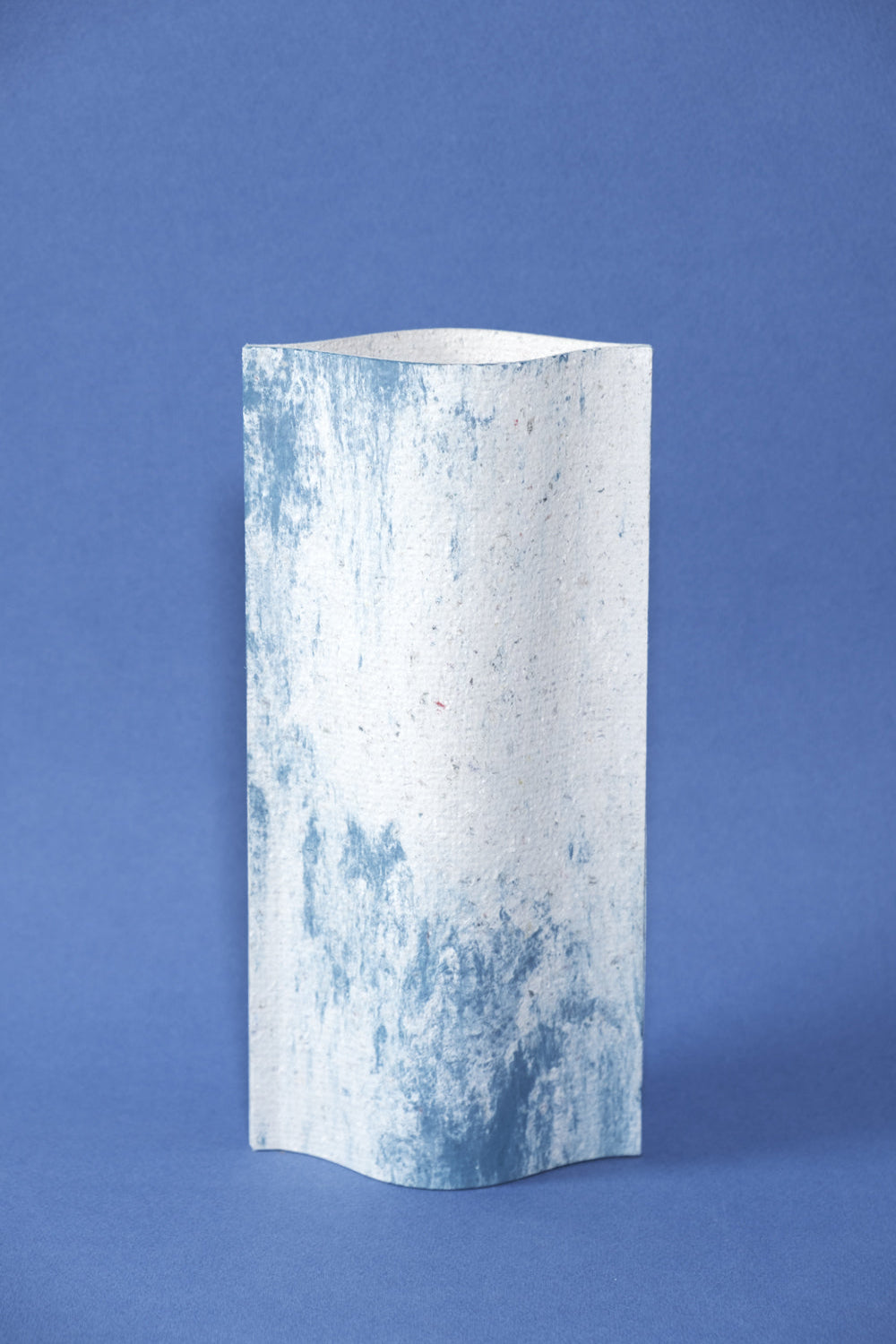 Un vase de profil rectangulaire sur fond bleu foncé. Deux feuilles de carton-pierre sont collées à leurs extrémités et ouvertes en leur centre pour y laisser passer un contenant étanche. Le vase arbore un effet marbré avec des veines couleur bleu marine sur fond blanc. Chaque vase a un marbré unique.