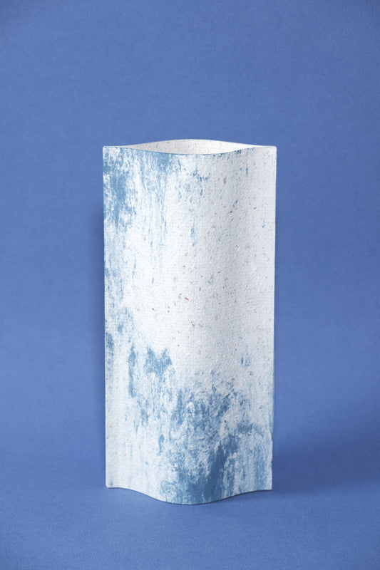 Un vase de profil rectangulaire sur fond bleu foncé. Deux feuilles de carton-pierre sont collées à leurs extrémités et ouvertes en leur centre pour y laisser passer un contenant étanche. Le vase arbore un effet marbré avec des veines couleur bleu marine sur fond blanc. Chaque vase a un marbré unique.