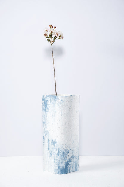 Un vase de profil rectangulaire sur fond blanc. Une branche y est soigneusement disposée. Deux feuilles de carton-pierre sont collées à leurs extrémités et ouvertes en leur centre pour y laisser passer un contenant étanche. Le vase arbore un effet marbré avec des veines couleur bleu sur fond blanc. Chaque vase a un marbré unique.