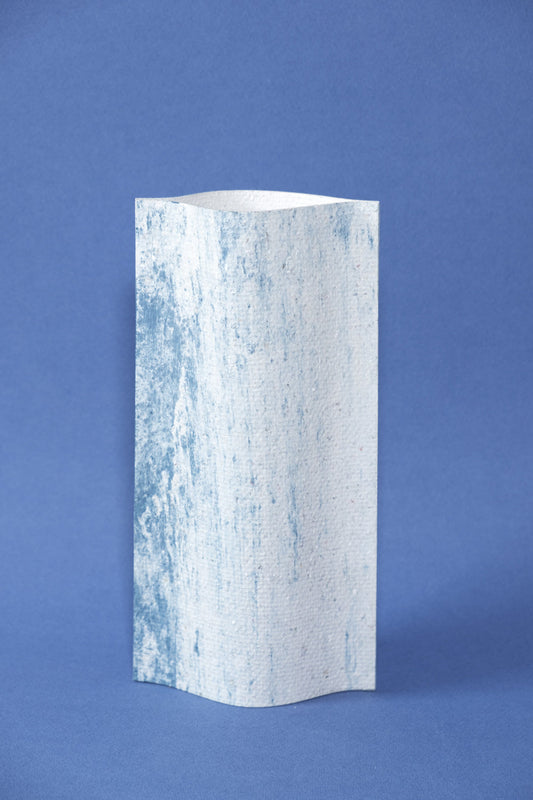 Un vase de profil rectangulaire sur fond bleu foncé. Deux feuilles de carton-pierre sont collées à leurs extrémités et ouvertes en leur centre pour y laisser passer un contenant étanche. Le vase arbore un effet marbré avec des veines couleur bleu marine sur fond blanc. Chaque vase a un marbré unique.