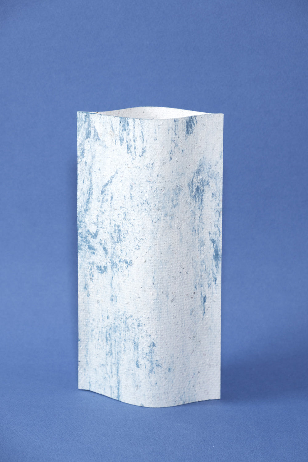 Un vase de profil rectangulaire sur fond bleu foncé. Deux feuilles de carton-pierre sont collées à leurs extrémités et ouvertes en leur centre pour y laisser passer un contenant étanche. Le vase arbore un effet marbré avec des veines couleur bleu marine sur fond blanc. Chaque vase a un marbré unique.