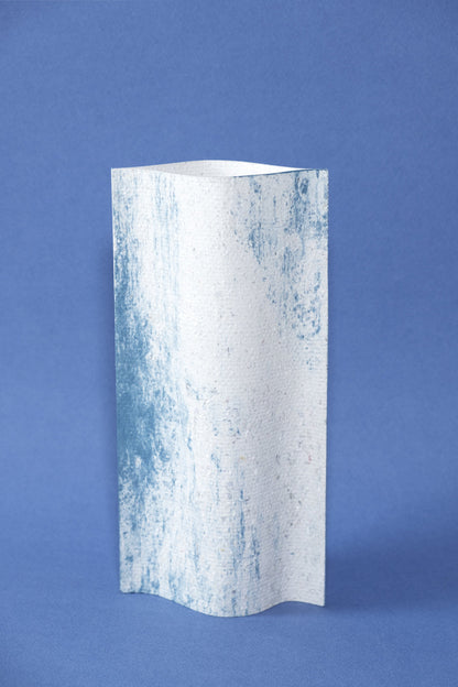 Un vase de profil rectangulaire sur fond bleu foncé. Deux feuilles de carton-pierre sont collées à leurs extrémités et ouvertes en leur centre pour y laisser passer un contenant étanche. Le vase arbore un effet marbré avec des veines couleur bleu marine sur fond blanc. Chaque vase a un marbré unique.