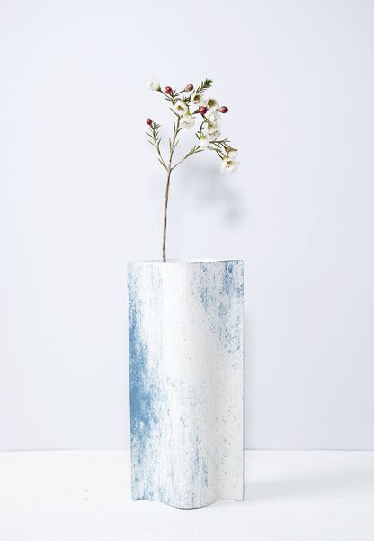 Un vase de profil rectangulaire sur fond blanc. Une branche y est soigneusement disposée. Deux feuilles de carton-pierre sont collées à leurs extrémités et ouvertes en leur centre pour y laisser passer un contenant étanche. Le vase arbore un effet marbré avec des veines couleur bleu sur fond blanc. Chaque vase a un marbré unique.