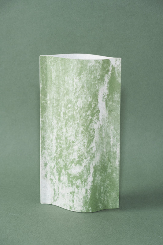 Un vase de profil rectangulaire sur fond vert foncé. Deux feuilles de carton-pierre sont collées à leurs extrémités et ouvertes en leur centre pour y laisser passer un contenant étanche. Le vase arbore un effet marbré avec des veines couleur vert tendre sur fond blanc. Chaque vase a un marbré unique.