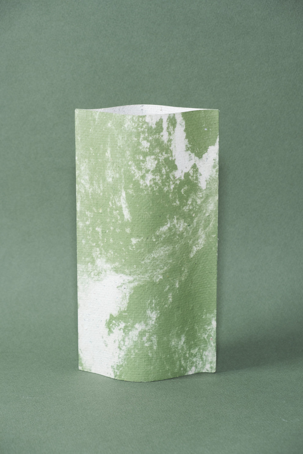 Un vase de profil rectangulaire sur fond vert foncé. Deux feuilles de carton-pierre sont collées à leurs extrémités et ouvertes en leur centre pour y laisser passer un contenant étanche. Le vase arbore un effet marbré avec des veines couleur vert tendre sur fond blanc. Chaque vase a un marbré unique.