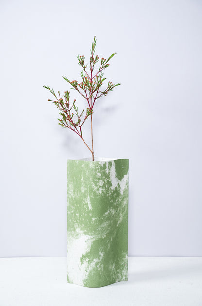 Un vase de profil rectangulaire sur fond blanc. Une branche y est soigneusement disposée. Deux feuilles de carton-pierre sont collées à leurs extrémités et ouvertes en leur centre pour y laisser passer un contenant étanche. Le vase arbore un effet marbré avec des veines couleur verte sur fond blanc. Chaque vase a un marbré unique.