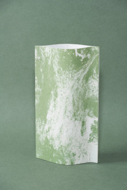 Un vase de profil rectangulaire sur fond vert foncé. Deux feuilles de carton-pierre sont collées à leurs extrémités et ouvertes en leur centre pour y laisser passer un contenant étanche. Le vase arbore un effet marbré avec des veines couleur vert tendre sur fond blanc. Chaque vase a un marbré unique.