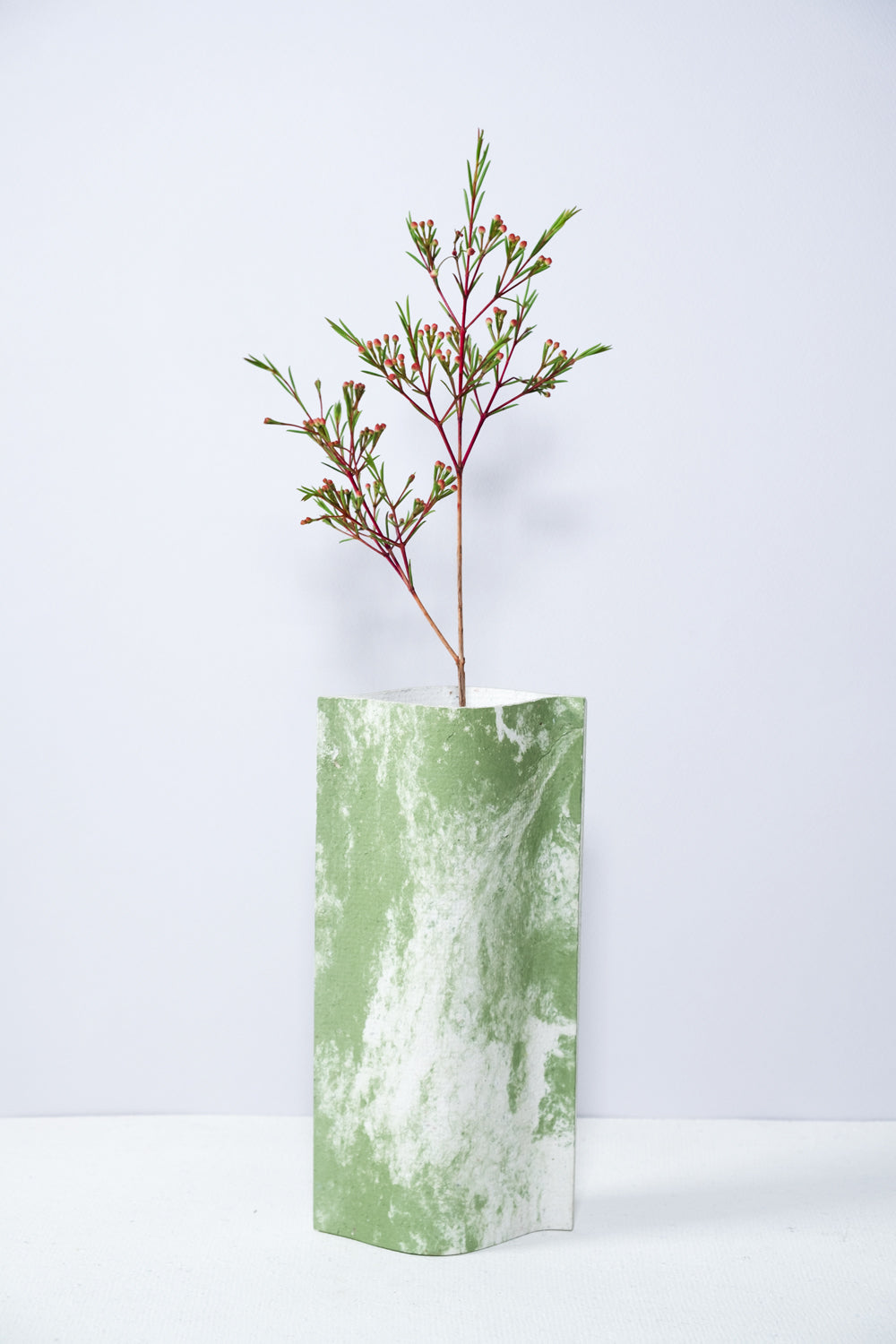 Un vase de profil rectangulaire sur fond blanc. Une branche y est soigneusement disposée. Deux feuilles de carton-pierre sont collées à leurs extrémités et ouvertes en leur centre pour y laisser passer un contenant étanche. Le vase arbore un effet marbré avec des veines couleur verte sur fond blanc. Chaque vase a un marbré unique.