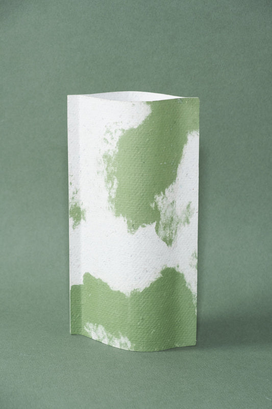 Un vase de profil rectangulaire sur fond vert foncé. Deux feuilles de carton-pierre sont collées à leurs extrémités et ouvertes en leur centre pour y laisser passer un contenant étanche. Le vase arbore un effet marbré avec des veines couleur vert tendre sur fond blanc. Chaque vase a un marbré unique.