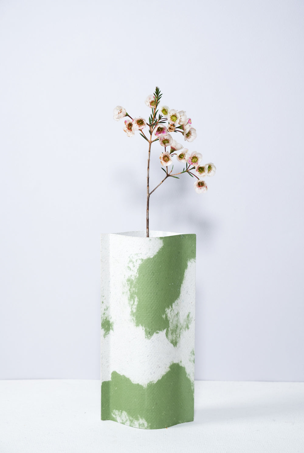 Un vase de profil rectangulaire sur fond blanc. Une branche y est soigneusement disposée. Deux feuilles de carton-pierre sont collées à leurs extrémités et ouvertes en leur centre pour y laisser passer un contenant étanche. Le vase arbore un effet marbré avec des veines couleur verte sur fond blanc. Chaque vase a un marbré unique.