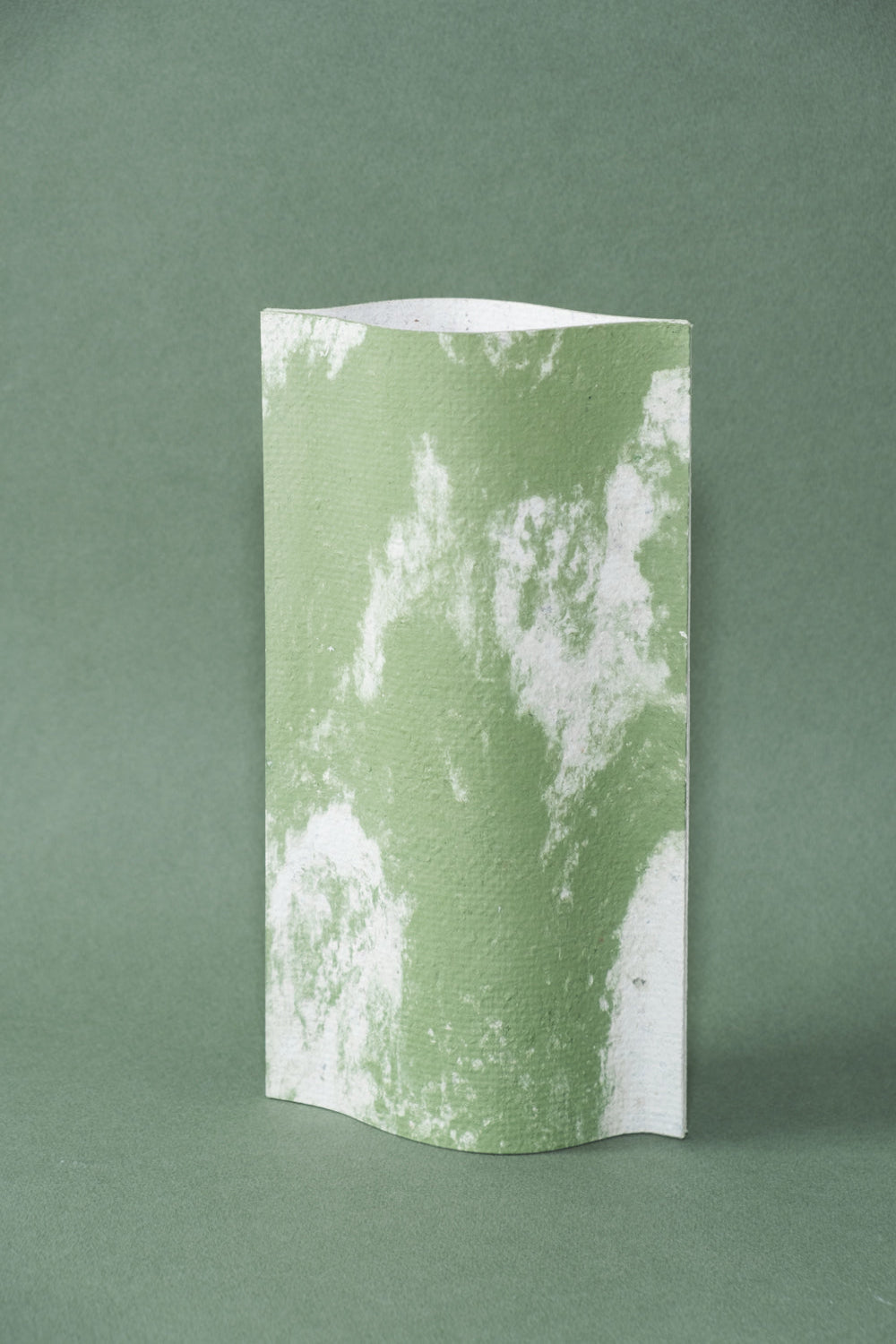 Un vase de profil rectangulaire sur fond vert foncé. Deux feuilles de carton-pierre sont collées à leurs extrémités et ouvertes en leur centre pour y laisser passer un contenant étanche. Le vase arbore un effet marbré avec des veines couleur vert tendre sur fond blanc. Chaque vase a un marbré unique.