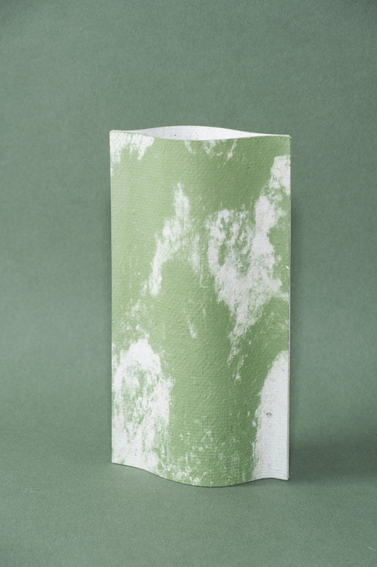 Un vase de profil rectangulaire sur fond vert foncé. Deux feuilles de carton-pierre sont collées à leurs extrémités et ouvertes en leur centre pour y laisser passer un contenant étanche. Le vase arbore un effet marbré avec des veines couleur vert tendre sur fond blanc. Chaque vase a un marbré unique.