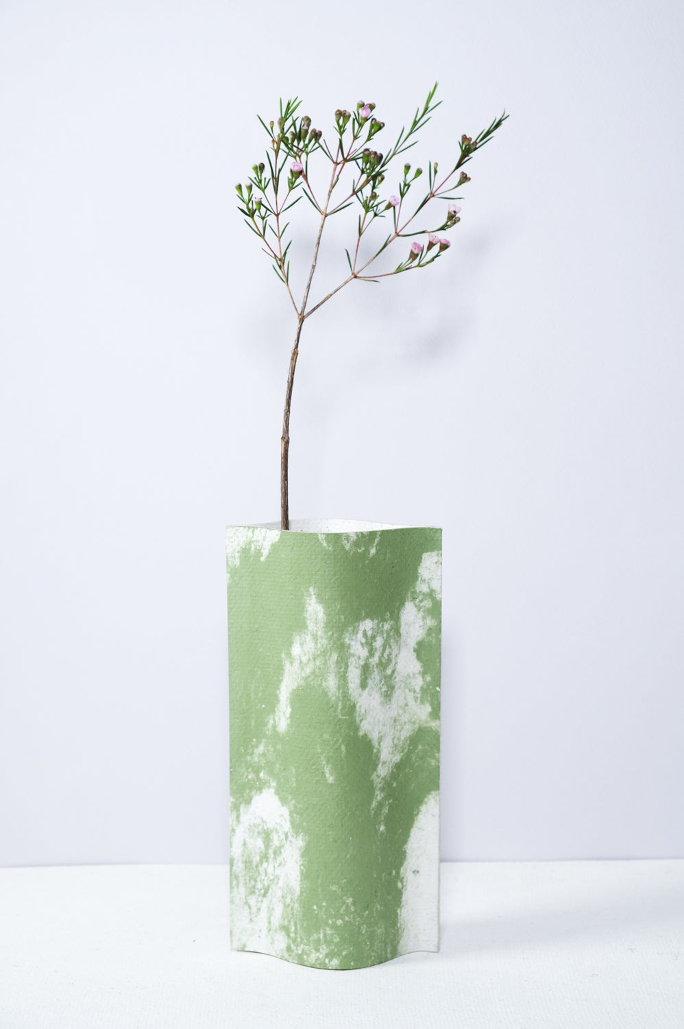 Un vase de profil rectangulaire sur fond blanc. Une branche y est soigneusement disposée. Deux feuilles de carton-pierre sont collées à leurs extrémités et ouvertes en leur centre pour y laisser passer un contenant étanche. Le vase arbore un effet marbré avec des veines couleur verte sur fond blanc. Chaque vase a un marbré unique.