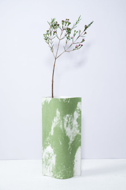 Un vase de profil rectangulaire sur fond blanc. Une branche y est soigneusement disposée. Deux feuilles de carton-pierre sont collées à leurs extrémités et ouvertes en leur centre pour y laisser passer un contenant étanche. Le vase arbore un effet marbré avec des veines couleur verte sur fond blanc. Chaque vase a un marbré unique.