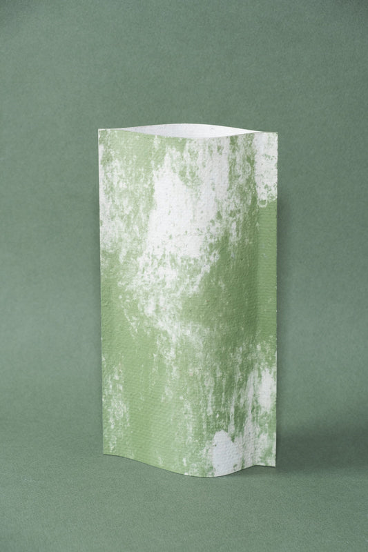 Un vase de profil rectangulaire sur fond vert foncé. Deux feuilles de carton-pierre sont collées à leurs extrémités et ouvertes en leur centre pour y laisser passer un contenant étanche. Le vase arbore un effet marbré avec des veines couleur vert tendre sur fond blanc. Chaque vase a un marbré unique.