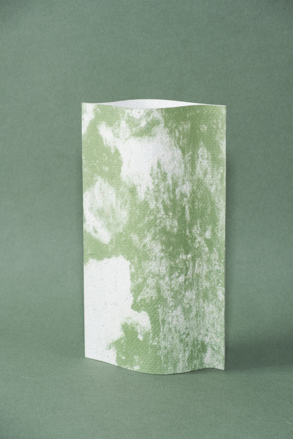 Un vase de profil rectangulaire sur fond vert foncé. Deux feuilles de carton-pierre sont collées à leurs extrémités et ouvertes en leur centre pour y laisser passer un contenant étanche. Le vase arbore un effet marbré avec des veines couleur vert tendre sur fond blanc. Chaque vase a un marbré unique.