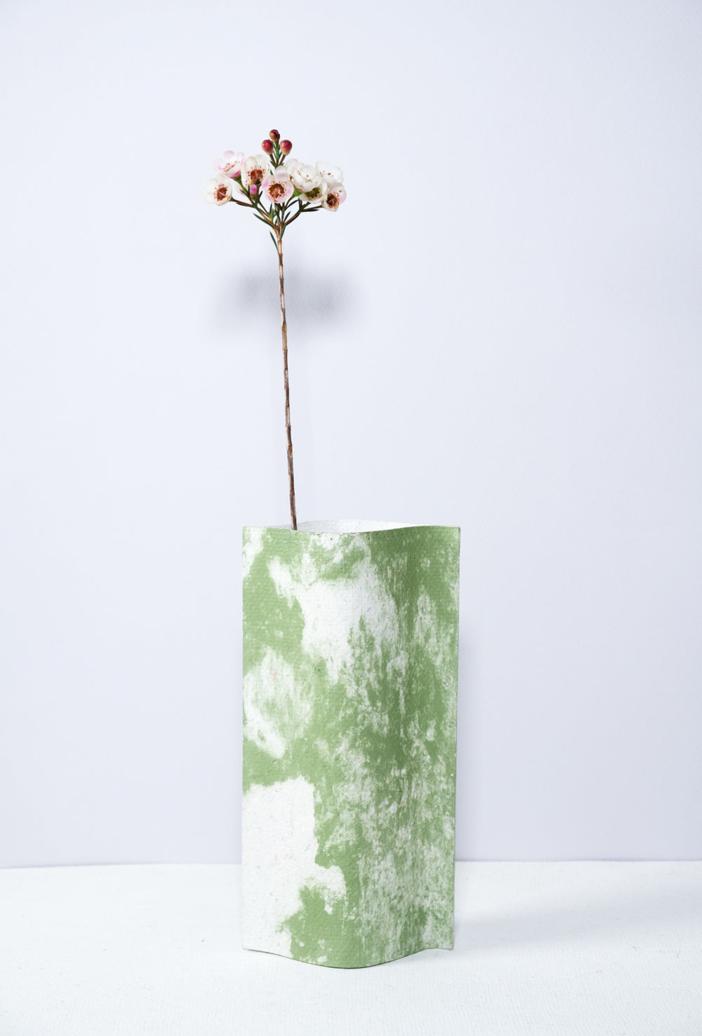 Un vase de profil rectangulaire sur fond blanc. Une branche y est soigneusement disposée. Deux feuilles de carton-pierre sont collées à leurs extrémités et ouvertes en leur centre pour y laisser passer un contenant étanche. Le vase arbore un effet marbré avec des veines couleur verte sur fond blanc. Chaque vase a un marbré unique.