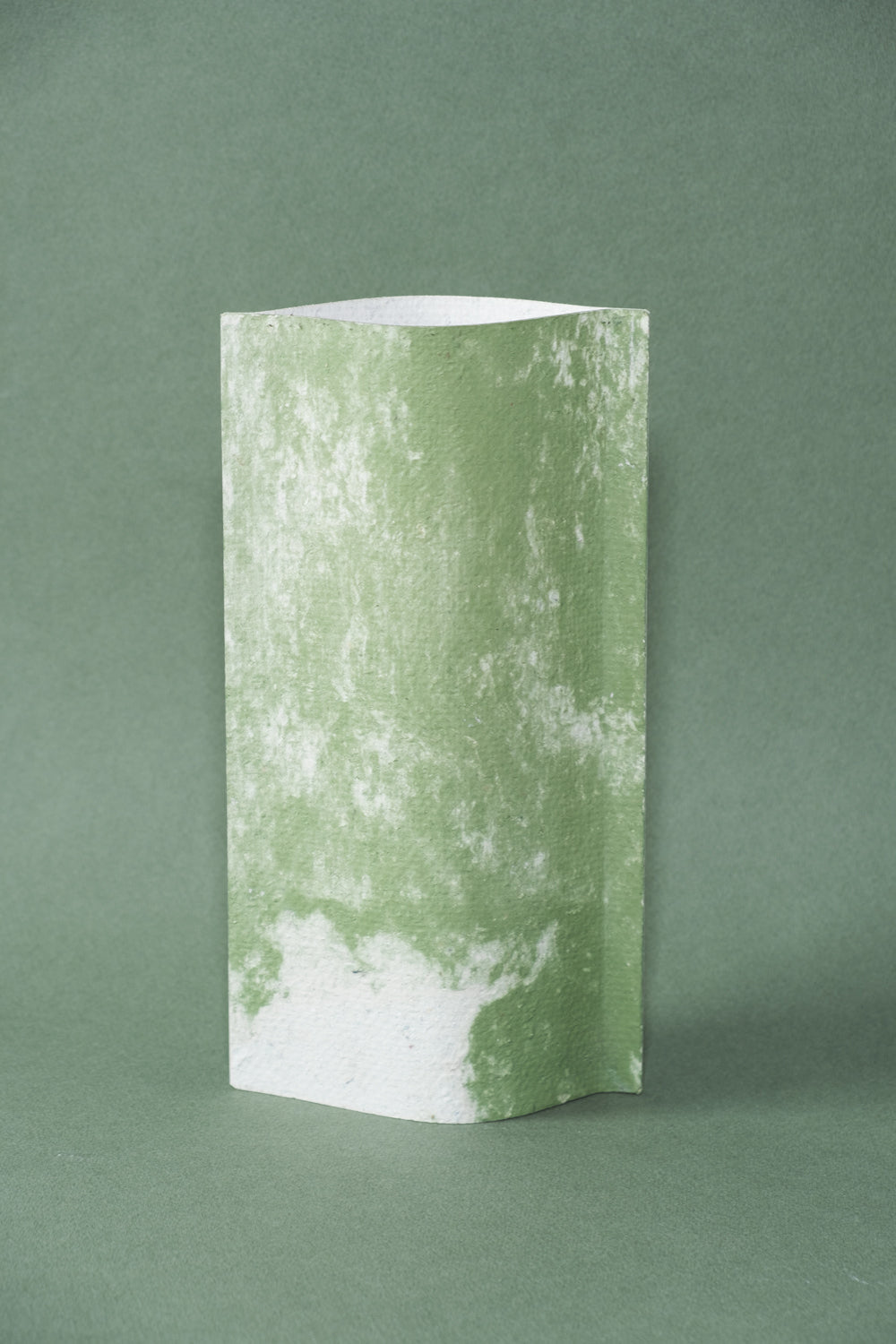 Un vase de profil rectangulaire sur fond vert foncé. Deux feuilles de carton-pierre sont collées à leurs extrémités et ouvertes en leur centre pour y laisser passer un contenant étanche. Le vase arbore un effet marbré avec des veines couleur vert tendre sur fond blanc. Chaque vase a un marbré unique.