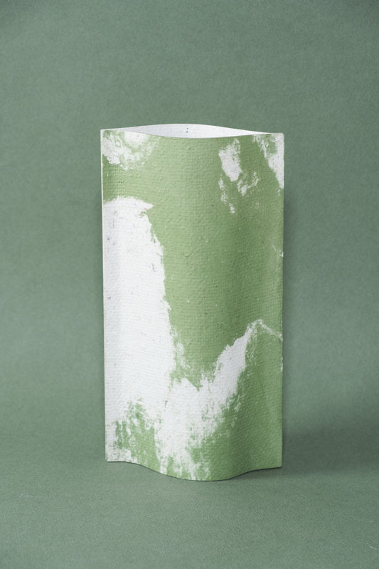 Un vase de profil rectangulaire sur fond vert foncé. Deux feuilles de carton-pierre sont collées à leurs extrémités et ouvertes en leur centre pour y laisser passer un contenant étanche. Le vase arbore un effet marbré avec des veines couleur vert tendre sur fond blanc. Chaque vase a un marbré unique.