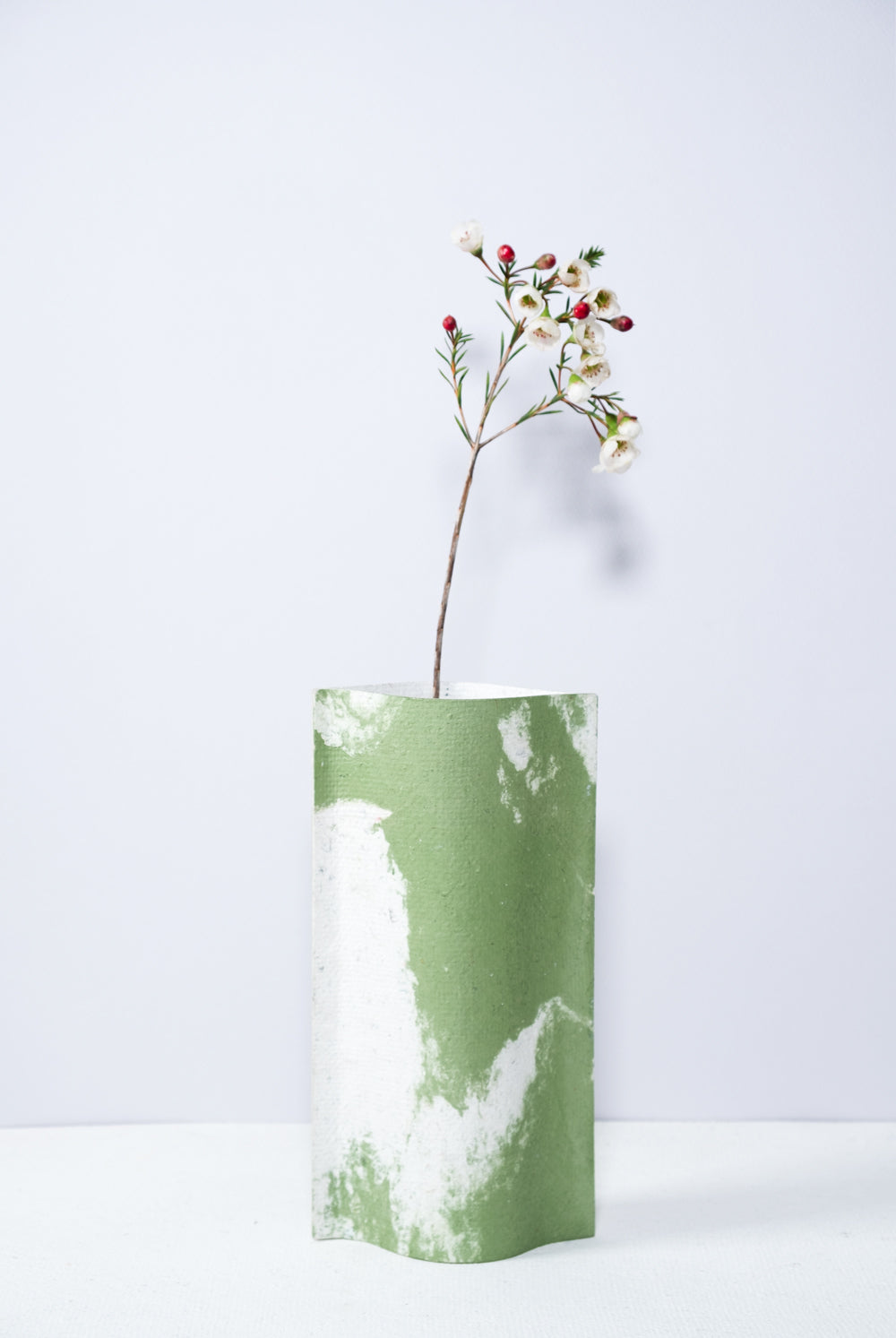 Un vase de profil rectangulaire sur fond blanc. Une branche y est soigneusement disposée. Deux feuilles de carton-pierre sont collées à leurs extrémités et ouvertes en leur centre pour y laisser passer un contenant étanche. Le vase arbore un effet marbré avec des veines couleur verte sur fond blanc. Chaque vase a un marbré unique.