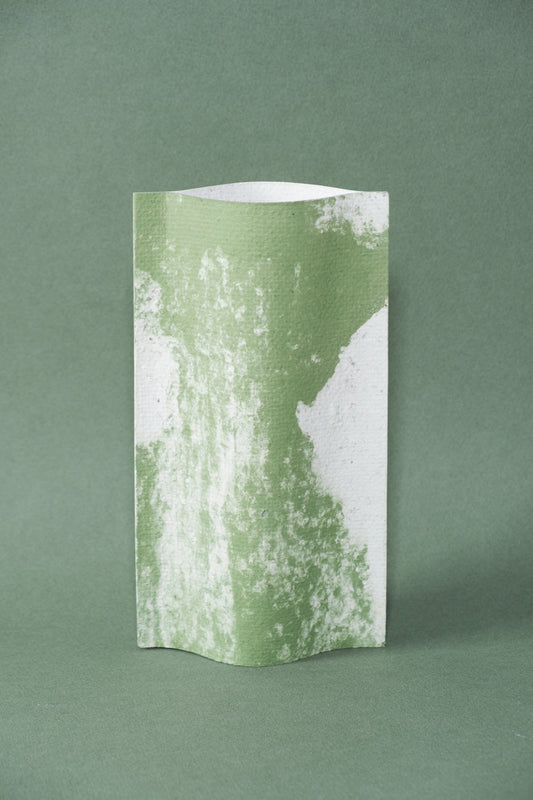 Un vase de profil rectangulaire sur fond vert foncé. Deux feuilles de carton-pierre sont collées à leurs extrémités et ouvertes en leur centre pour y laisser passer un contenant étanche. Le vase arbore un effet marbré avec des veines couleur vert tendre sur fond blanc. Chaque vase a un marbré unique.