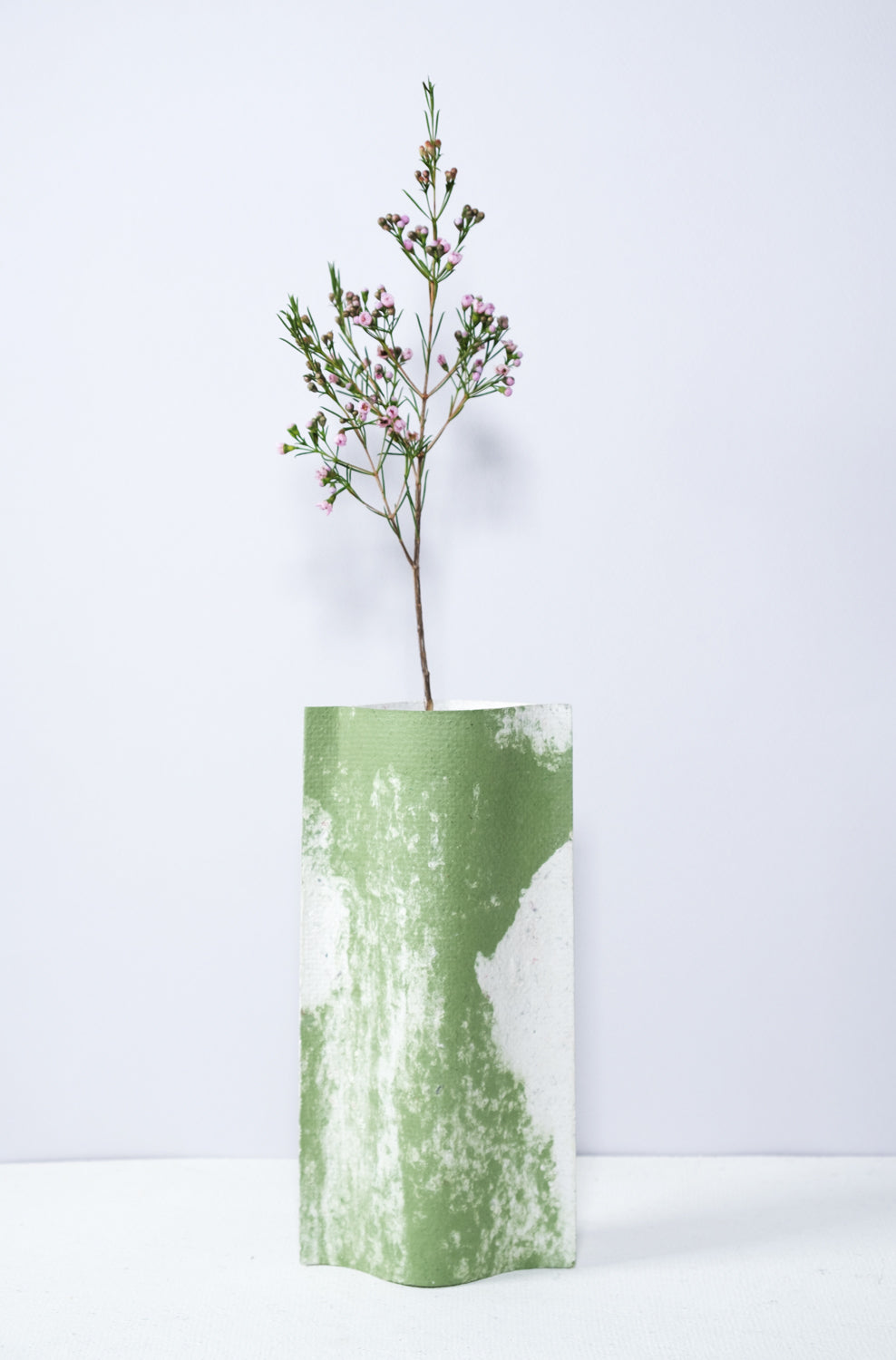 Un vase de profil rectangulaire sur fond blanc. Une branche y est soigneusement disposée. Deux feuilles de carton-pierre sont collées à leurs extrémités et ouvertes en leur centre pour y laisser passer un contenant étanche. Le vase arbore un effet marbré avec des veines couleur verte sur fond blanc. Chaque vase a un marbré unique.