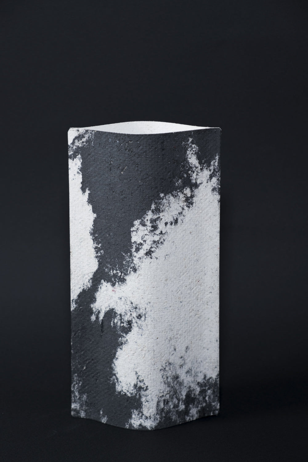 Un vase de profil rectangulaire sur fond noir. Deux feuilles de carton-pierre sont collées à leurs extrémités et ouvertes en leur centre pour y laisser passer un contenant étanche. Le vase arbore un effet marbré avec des veines couleur noir charbon sur fond blanc. Chaque vase a un marbré unique.