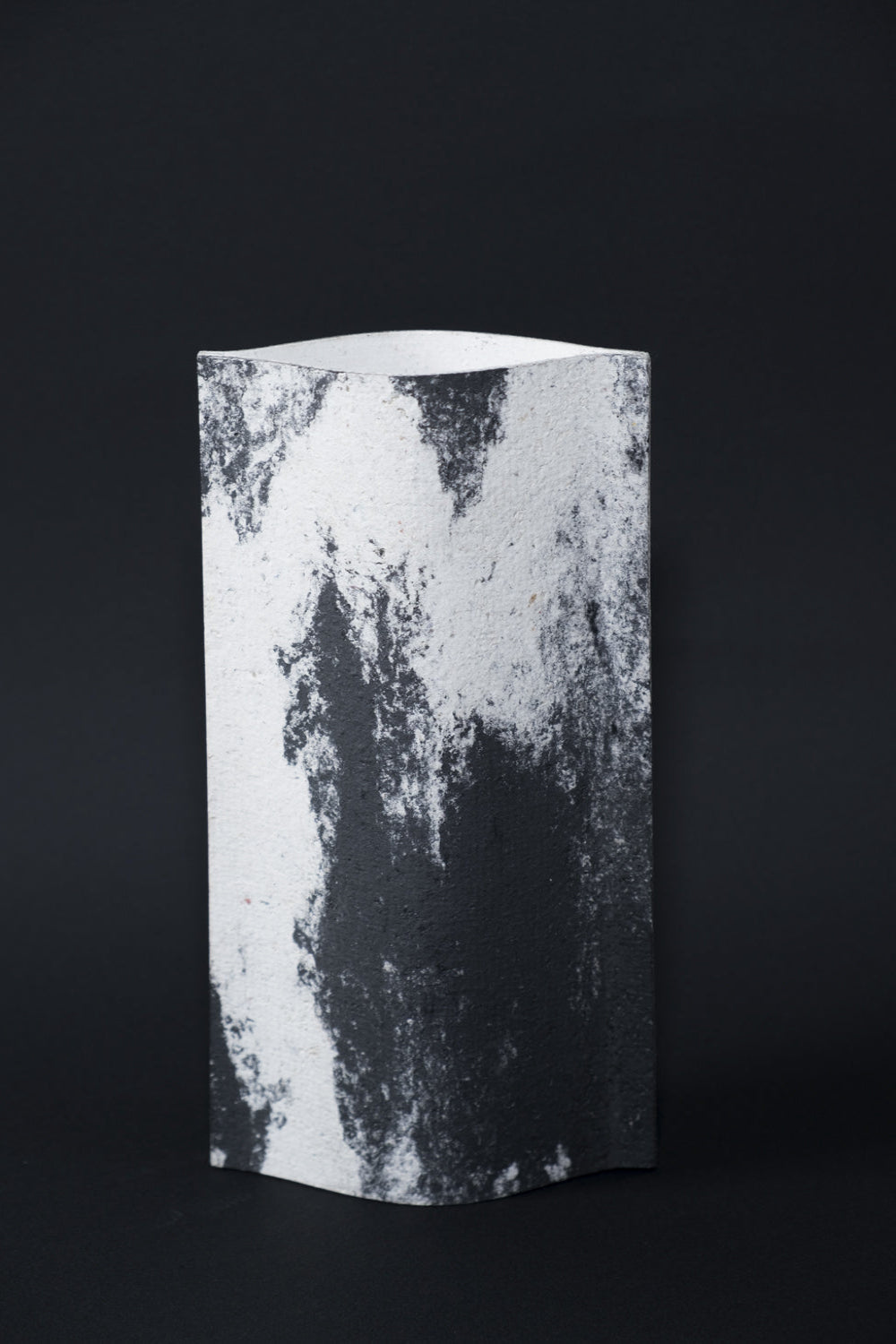 Un vase de profil rectangulaire sur fond noir. Deux feuilles de carton-pierre sont collées à leurs extrémités et ouvertes en leur centre pour y laisser passer un contenant étanche. Le vase arbore un effet marbré avec des veines couleur noir charbon sur fond blanc. Chaque vase a un marbré unique.