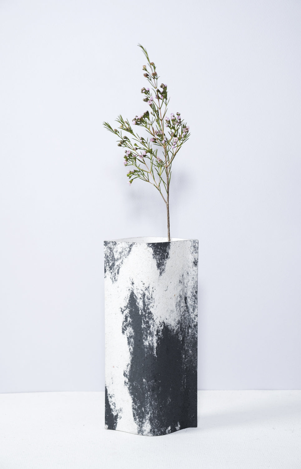 Un vase de profil rectangulaire sur fond blanc. Une branche y est soigneusement disposée. Deux feuilles de carton-pierre sont collées à leurs extrémités et ouvertes en leur centre pour y laisser passer un contenant étanche. Le vase arbore un effet marbré avec des veines couleur noir sur fond blanc. Chaque vase a un marbré unique.