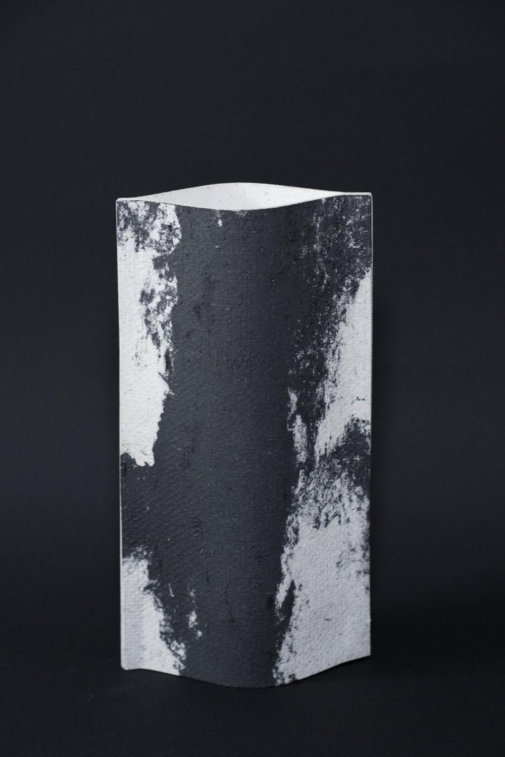 Un vase de profil rectangulaire sur fond noir. Deux feuilles de carton-pierre sont collées à leurs extrémités et ouvertes en leur centre pour y laisser passer un contenant étanche. Le vase arbore un effet marbré avec des veines couleur noir charbon sur fond blanc. Chaque vase a un marbré unique.