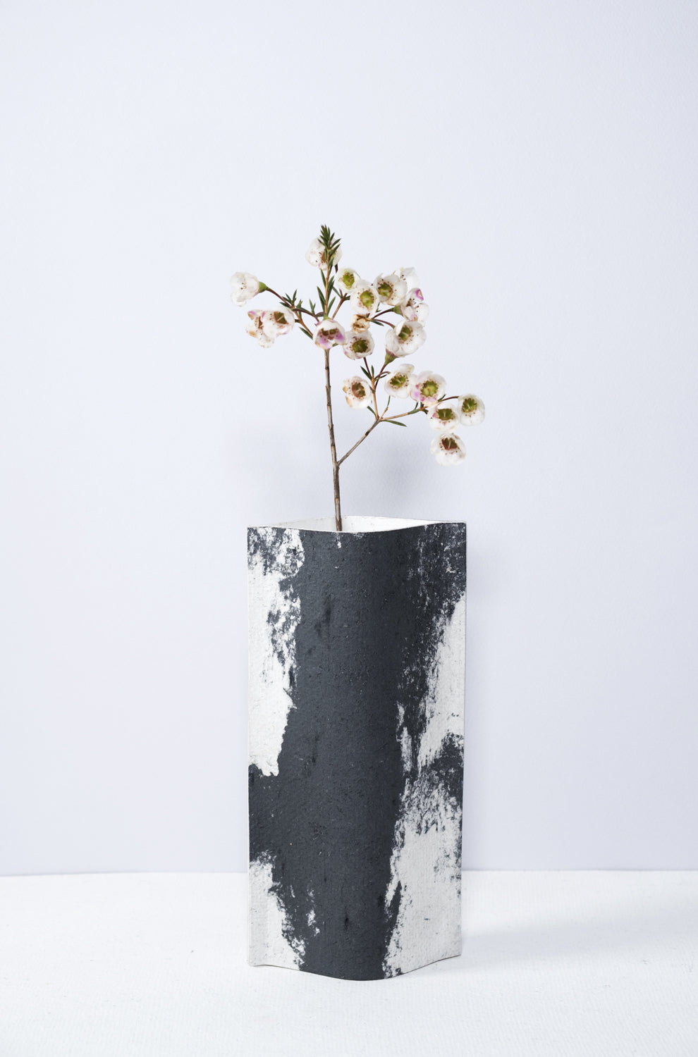 Un vase de profil rectangulaire sur fond blanc. Une branche y est soigneusement disposée. Deux feuilles de carton-pierre sont collées à leurs extrémités et ouvertes en leur centre pour y laisser passer un contenant étanche. Le vase arbore un effet marbré avec des veines couleur noir sur fond blanc. Chaque vase a un marbré unique.