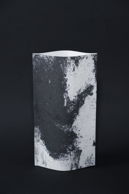 Un vase de profil rectangulaire sur fond noir. Deux feuilles de carton-pierre sont collées à leurs extrémités et ouvertes en leur centre pour y laisser passer un contenant étanche. Le vase arbore un effet marbré avec des veines couleur noir charbon sur fond blanc. Chaque vase a un marbré unique.