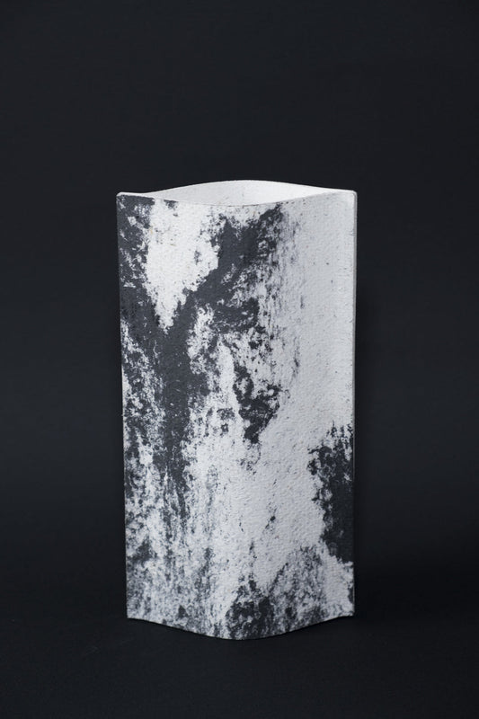 Un vase de profil rectangulaire sur fond noir. Deux feuilles de carton-pierre sont collées à leurs extrémités et ouvertes en leur centre pour y laisser passer un contenant étanche. Le vase arbore un effet marbré avec des veines couleur noir charbon sur fond blanc. Chaque vase a un marbré unique.