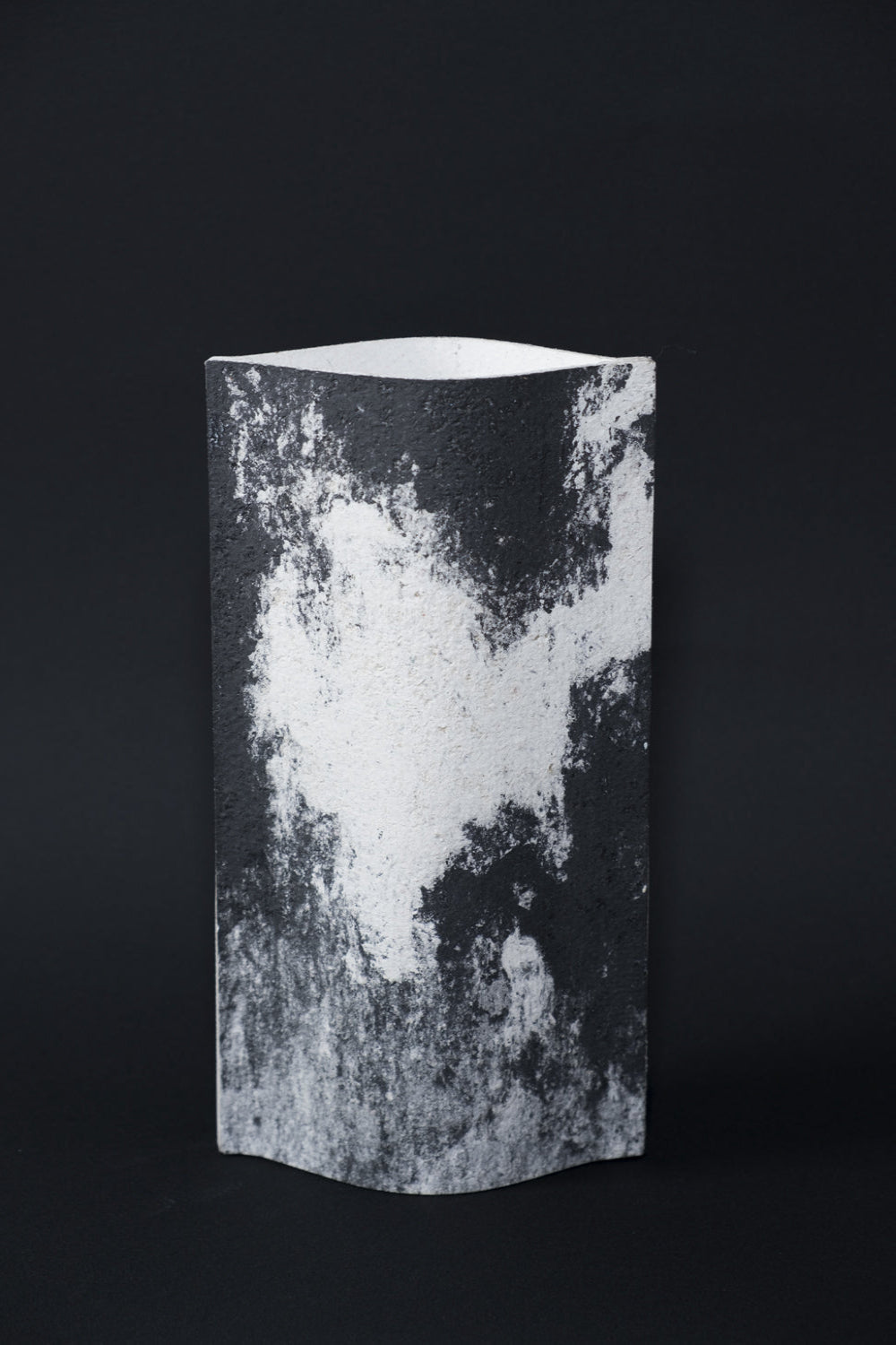 Un vase de profil rectangulaire sur fond noir. Deux feuilles de carton-pierre sont collées à leurs extrémités et ouvertes en leur centre pour y laisser passer un contenant étanche. Le vase arbore un effet marbré avec des veines couleur noir charbon sur fond blanc. Chaque vase a un marbré unique.