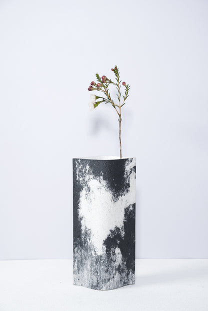 Un vase de profil rectangulaire sur fond blanc. Une branche y est soigneusement disposée. Deux feuilles de carton-pierre sont collées à leurs extrémités et ouvertes en leur centre pour y laisser passer un contenant étanche. Le vase arbore un effet marbré avec des veines couleur noir sur fond blanc. Chaque vase a un marbré unique.