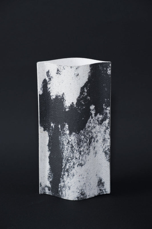 Un vase de profil rectangulaire sur fond noir. Deux feuilles de carton-pierre sont collées à leurs extrémités et ouvertes en leur centre pour y laisser passer un contenant étanche. Le vase arbore un effet marbré avec des veines couleur noir charbon sur fond blanc. Chaque vase a un marbré unique.