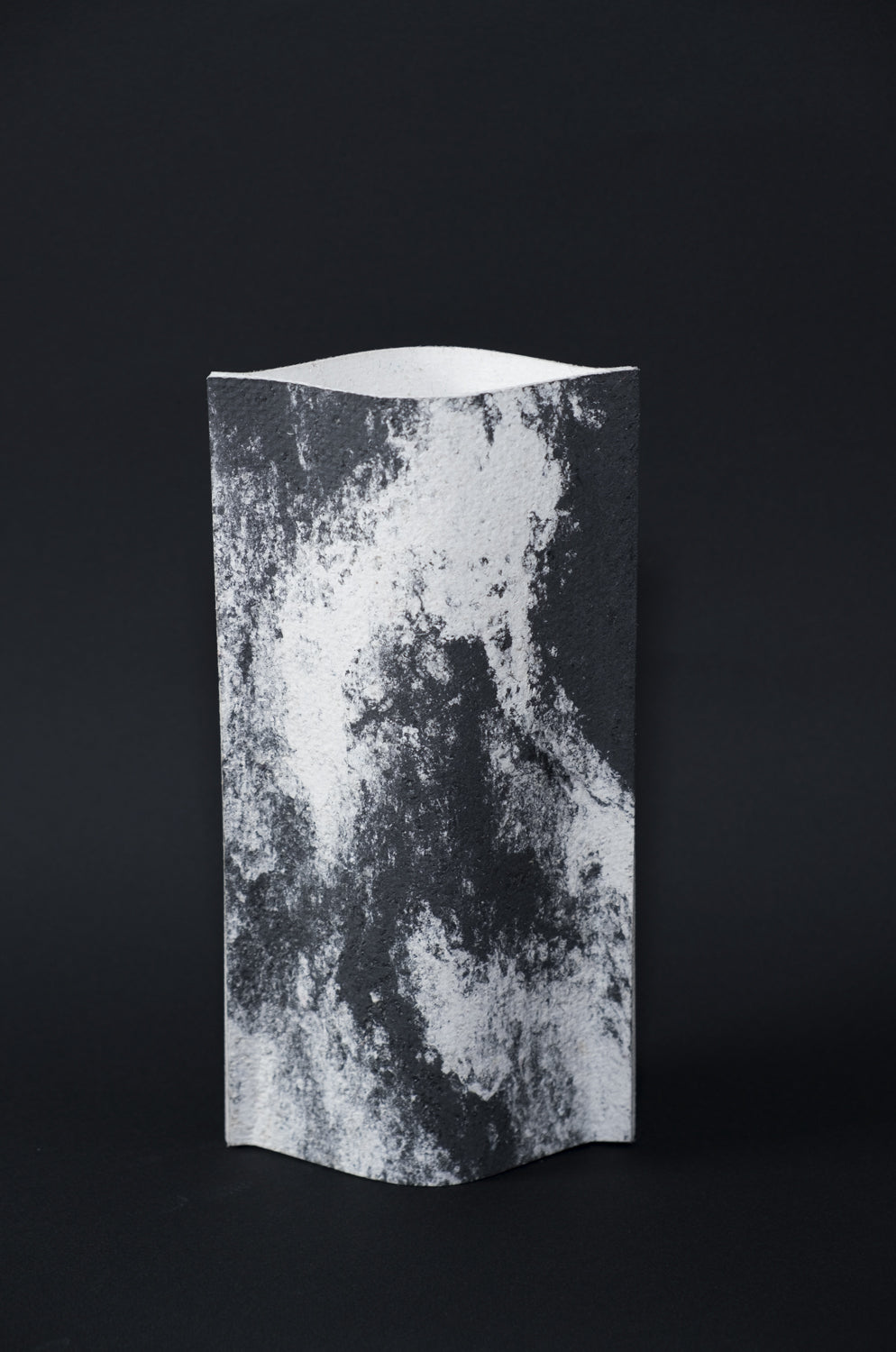 Un vase de profil rectangulaire sur fond noir. Deux feuilles de carton-pierre sont collées à leurs extrémités et ouvertes en leur centre pour y laisser passer un contenant étanche. Le vase arbore un effet marbré avec des veines couleur noir charbon sur fond blanc. Chaque vase a un marbré unique.