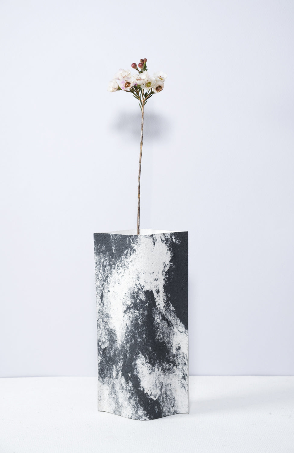 Un vase de profil rectangulaire sur fond blanc. Une branche y est soigneusement disposée. Deux feuilles de carton-pierre sont collées à leurs extrémités et ouvertes en leur centre pour y laisser passer un contenant étanche. Le vase arbore un effet marbré avec des veines couleur noir sur fond blanc. Chaque vase a un marbré unique.