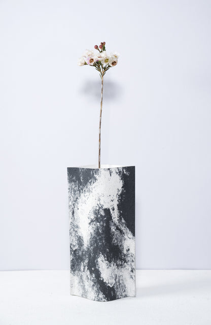 Un vase de profil rectangulaire sur fond blanc. Une branche y est soigneusement disposée. Deux feuilles de carton-pierre sont collées à leurs extrémités et ouvertes en leur centre pour y laisser passer un contenant étanche. Le vase arbore un effet marbré avec des veines couleur noir sur fond blanc. Chaque vase a un marbré unique.