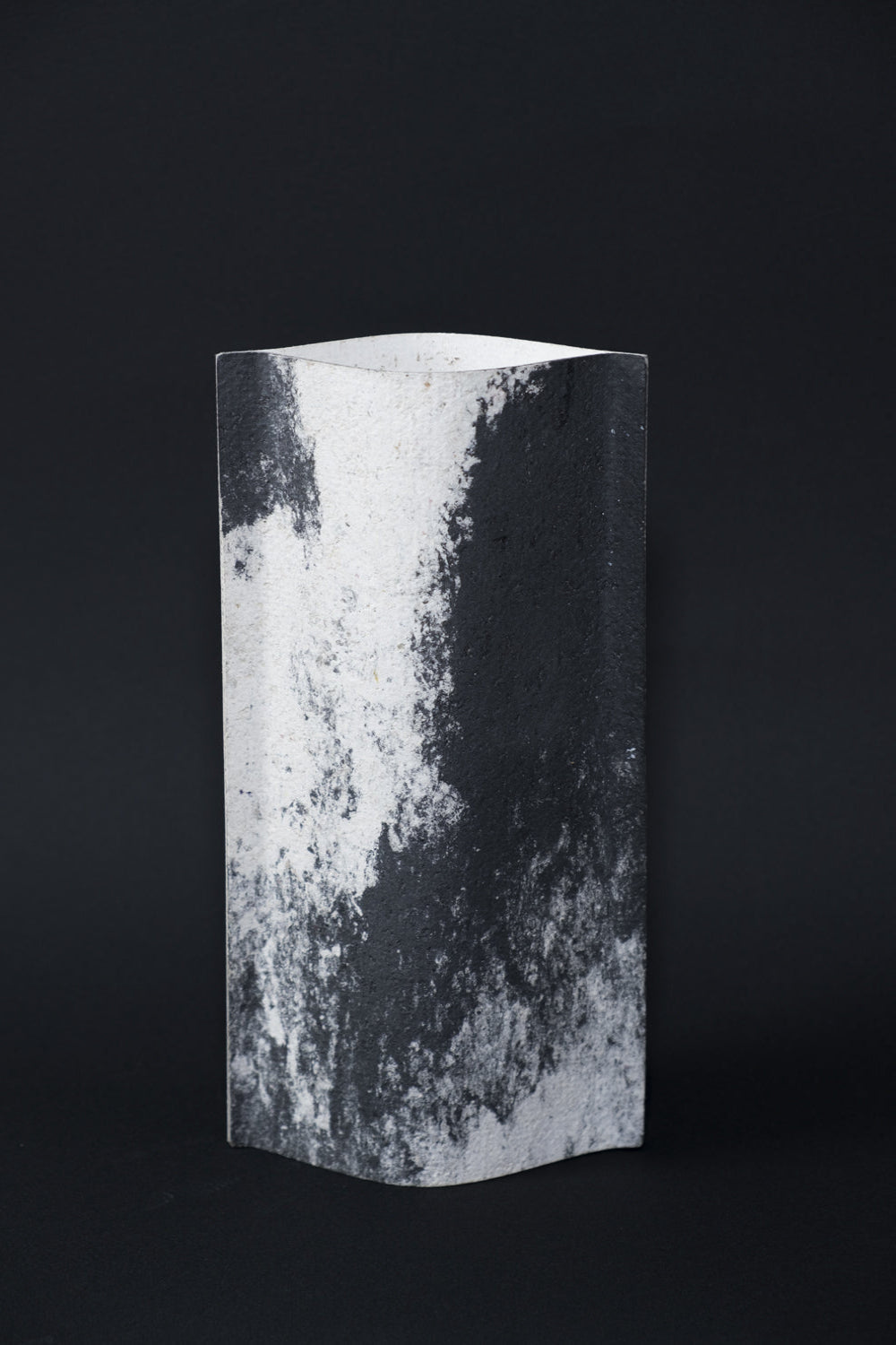 Un vase de profil rectangulaire sur fond noir. Deux feuilles de carton-pierre sont collées à leurs extrémités et ouvertes en leur centre pour y laisser passer un contenant étanche. Le vase arbore un effet marbré avec des veines couleur noir charbon sur fond blanc. Chaque vase a un marbré unique.