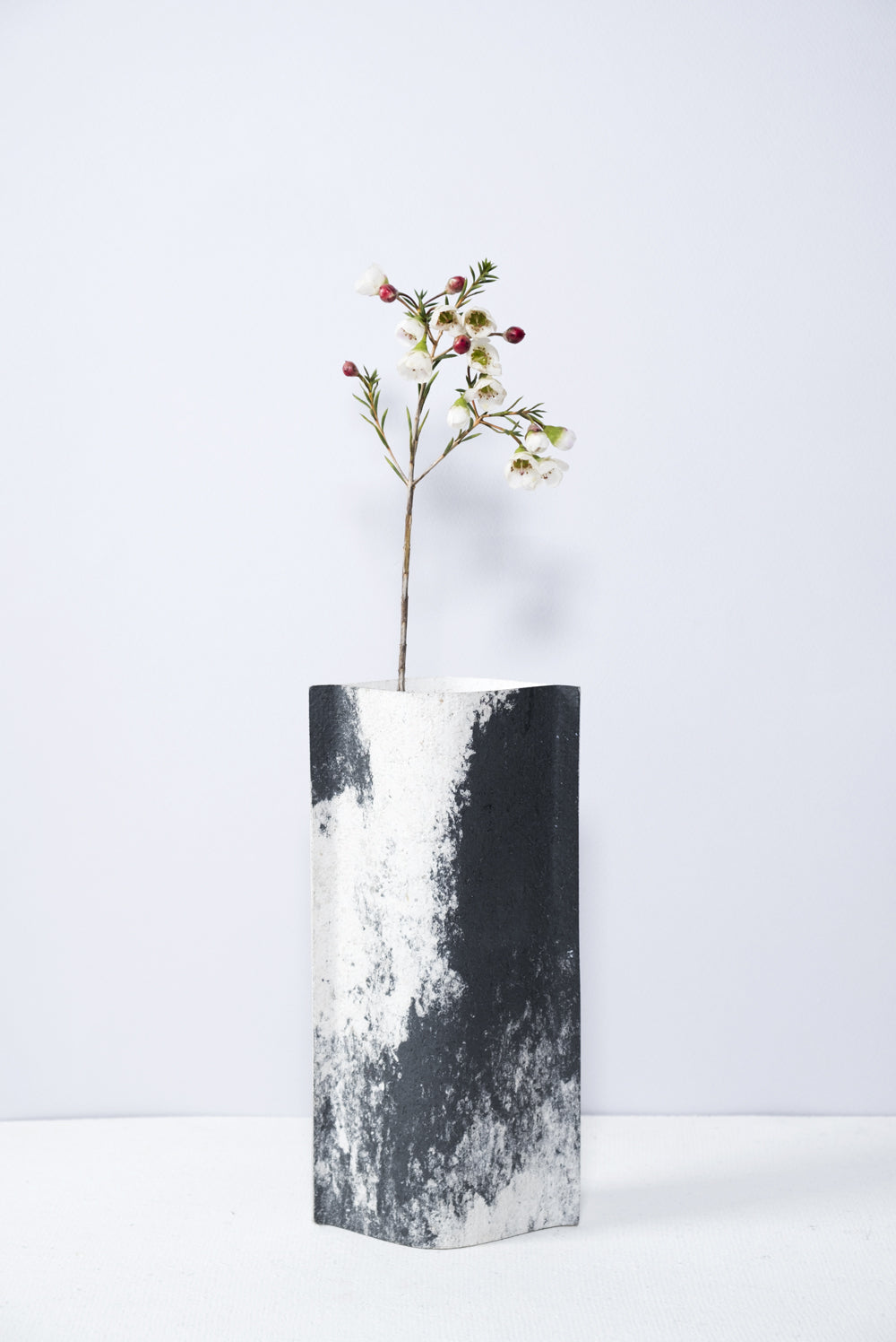 Un vase de profil rectangulaire sur fond blanc. Une branche y est soigneusement disposée. Deux feuilles de carton-pierre sont collées à leurs extrémités et ouvertes en leur centre pour y laisser passer un contenant étanche. Le vase arbore un effet marbré avec des veines couleur noir sur fond blanc. Chaque vase a un marbré unique.